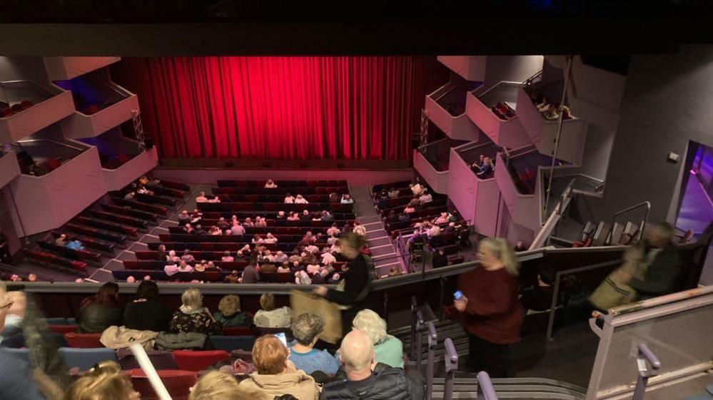 The circle and stalls of the Derngate, with audience members waiting for the red stage curtain to open. Pink-coloured boxes are visible on both sides.