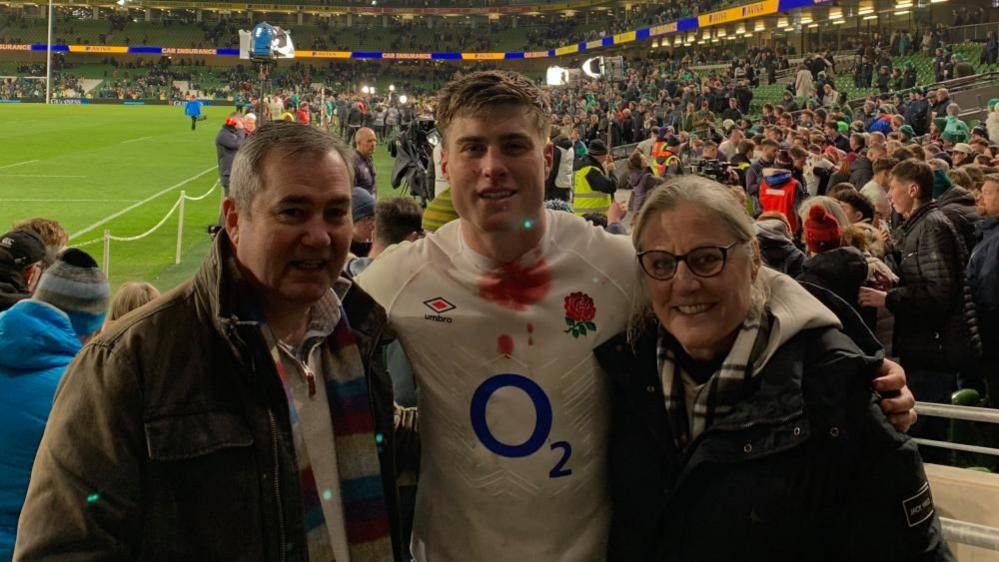 Cliff Freeman with short grey hair wearing a brown jacket, Tommy Freeman with short dark hair wearing a white England shirt with an O2 logo on the front, and Sara Freeman with short light-coloured hair and glasses wearing a dark jacket with white hood. Tommy has his arms around them and they are standing in a sports stadium with a crowd of people behind them. The rugby pitch is visible to the left.