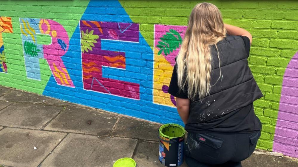 A closer-up view of a vibrant mural, painted along a low brick wall along a riverside path. The mural depicts the word 'Rowntree' painted in bright multicolours against predominately lime-green background. Each letter also contains leaf and flower motifs within it. In the corner, a woman with long blonde hair and wearing black paints onto the mural.