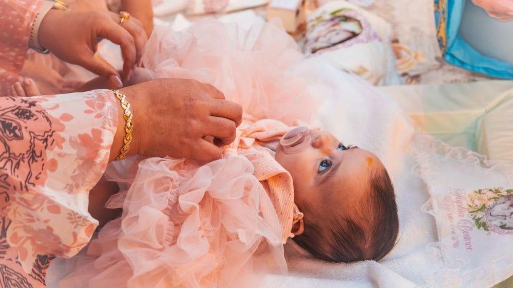 In this photograph baby Sophia is picture lying down wearing a soft pink ruffled dress and sucking on a dummy. She has tufts of brown hair and large dark eyes.