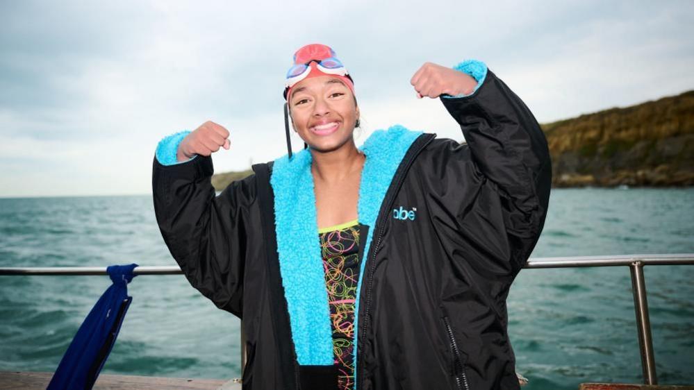 Prisha Tapre in a DryRobe and with her swimming goggles and swim hat on, standing on a boat and smiling with her arms in a strongman pose