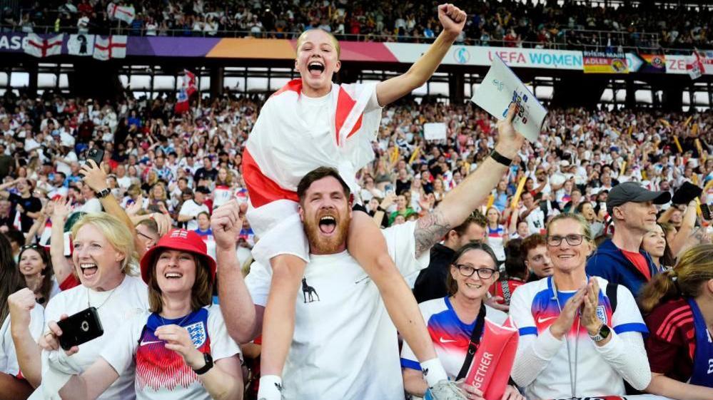 A young girl draped in an England flag sits on a man's shoulder surrounded by cheering England fans in a packed stadium