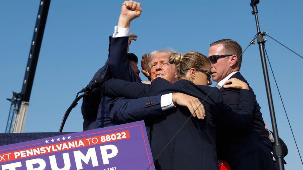 Donald Trump, surrounded by Secret Service agents wearing dark glasses who hold him close to them, raising a fist in defiance with a streak of blood across his face