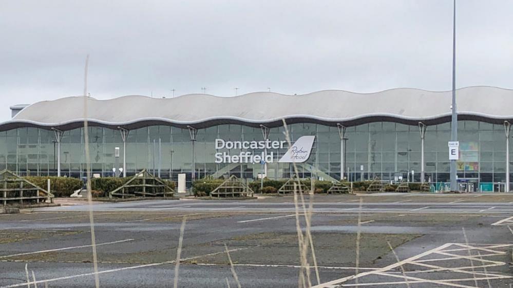 Exterior of Doncaster Sheffield Airport. It has a wavy roof and the front is fully glass. The car park in the foreground is empty.