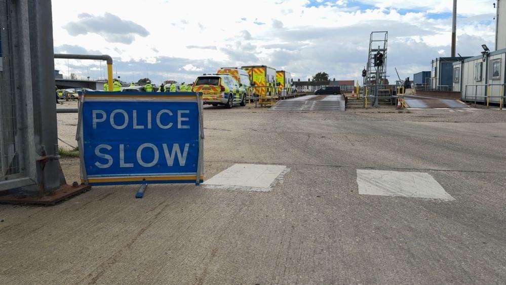 A "police slow" sign in white text with blue background. Beyond is a pair of weighbridges, and police and other agency vehicles lined up on hardstanding.
