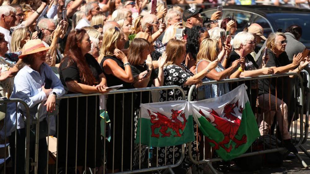 A large group of people are standing behind safety barriers, with two Welsh flags attached to the barriers. Many people are filming with their mobile phones.