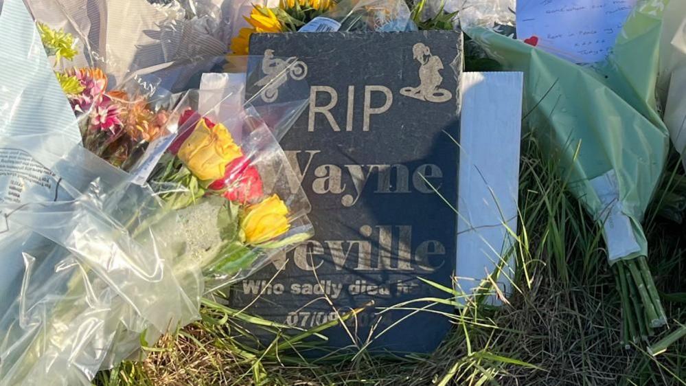 Close-up on a small slate sign, reading RIP Wayne Neville, who sadly died here 07/09/24. Surrounding it are bunches of red and yellow roses, and there's grass on the ground. It's lit up by sunlight. 