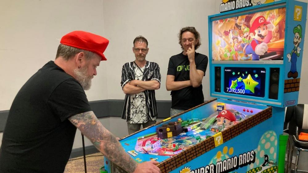 Man with dark hair wearing red cap plays a mainly blue machine with Super Mario graphics while two men watch