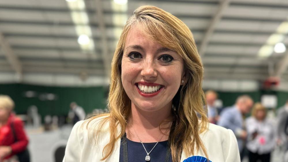 Sarah Bool with long blond hair wearing a cream jacket and blue top.  An election count is underway in the background.