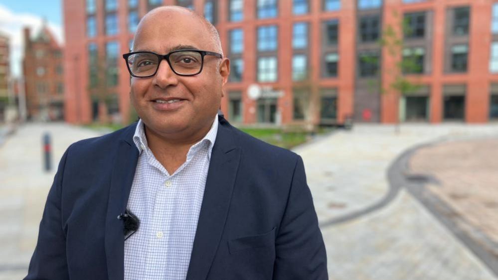 Baggy Shanker, in a suit jacket and open collar shirt and black glasses, smiles at the camera. There is a red brick office building in the background.