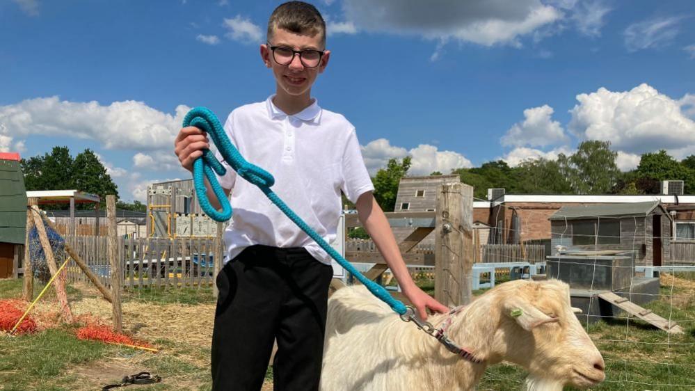 Kai, holding the reigns to a goat, in school grounds, with equipment behind him.