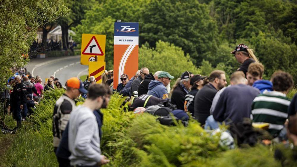 Spectators lining the hedge at Hillberry on the TT course. A sign for the milestone can be seen alongside other road signs, as the road sweeps up to the right.