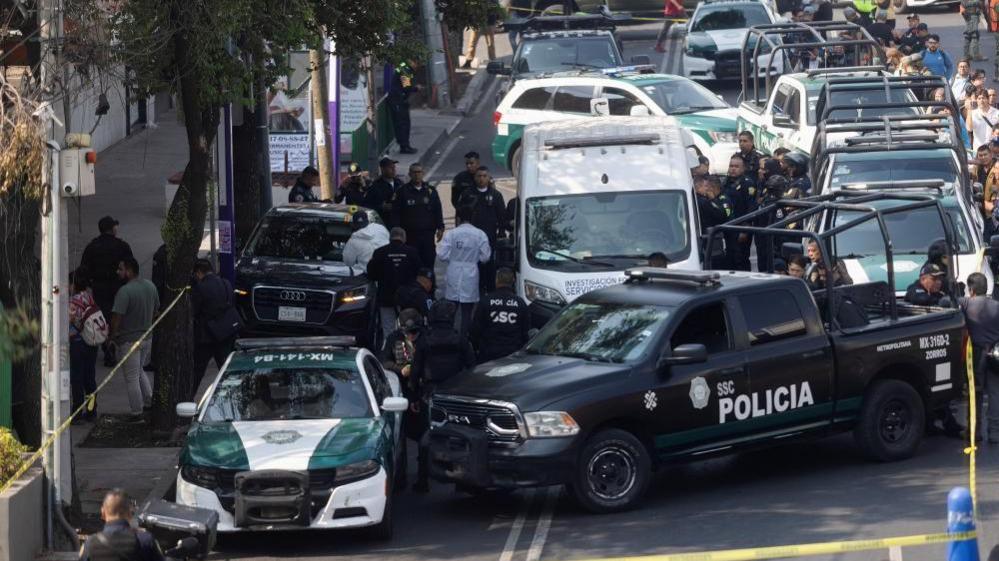 A black Audi with bullet holes in its windscreen is surrounded by police cars and SUVs. Crime scene tape has been strung around the cars and a man in a lab coat can be seen next to the car.