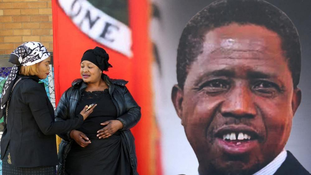 Mourners gather in front of a banner ahead of the funeral of Zambia's former President Edgar Lungu - the banner features a portrait of Lungu.