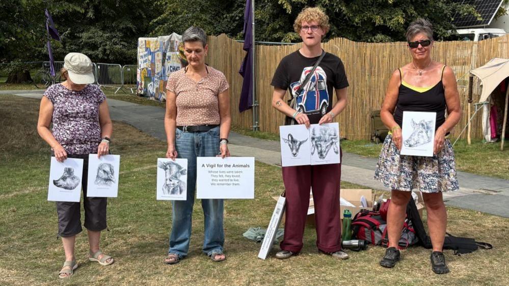 Four people stand in a line on grass. One is wearing a beige cap. They are holding drawings of animals and a sign which says "A vigil for the animals whose lives were taken". There is a path and a wooden fence behind them.