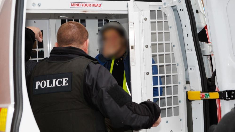 A police officer speaks to a man sitting in the back of a police van. The man's face is blurred. 