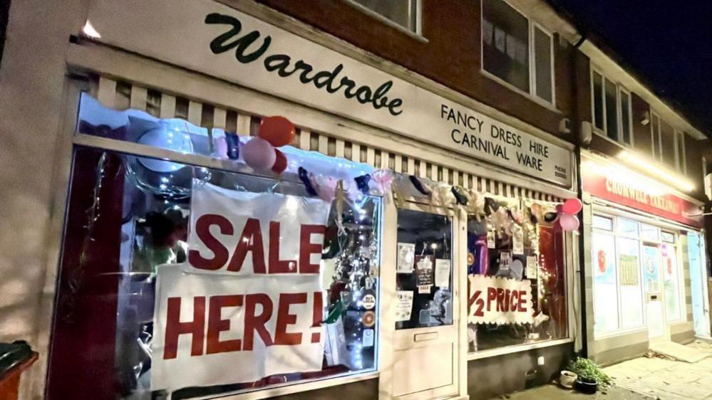 The Wardrobe shop front. A sign said "Wardrobe fancy dress hire, carnival ware". Red and pink balloons hang from the shop. Signs written in red capital letters say, "Sale Here" and "1/2 Price" 