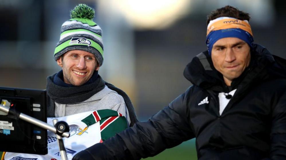 Sinfield embraces his friend Burrow at the end of a fundraising challenge. Sinfield wears his fundraising head band and a black thermal jacket, Burrow is wrapped in a woolly hat, scarf and blanket and smiles. Burrow's computer, which aided his speech, can be seen in the shot.