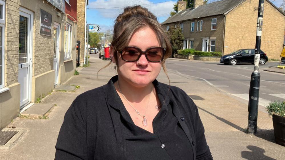 Lily Brown is standing on the high street in Lakenheath. You can see the road and a zebra crossing in the background.  Lily is wearing a black barbers top, with back cardigan. She has her hair up in a messy bun and is wearing sunglasses. 