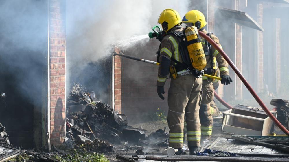 A firefighter in full breathing apparatus and a yellow helmet holds a piece of metal sheeting, which is in the middle of debris outside a row of burned-out garages. Another firefighter is standing in the entrance to another garage just behind him. There is a haze of smoke in the air around them.