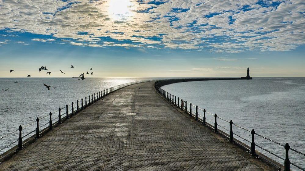 Roker Pier which is curved with railings on either side. In the distance is the lighthouse which is just a silhouette. A flock of birds are flying close to the pier on the left. The sky is blue and white with small, wispy clouds in front of the sun.