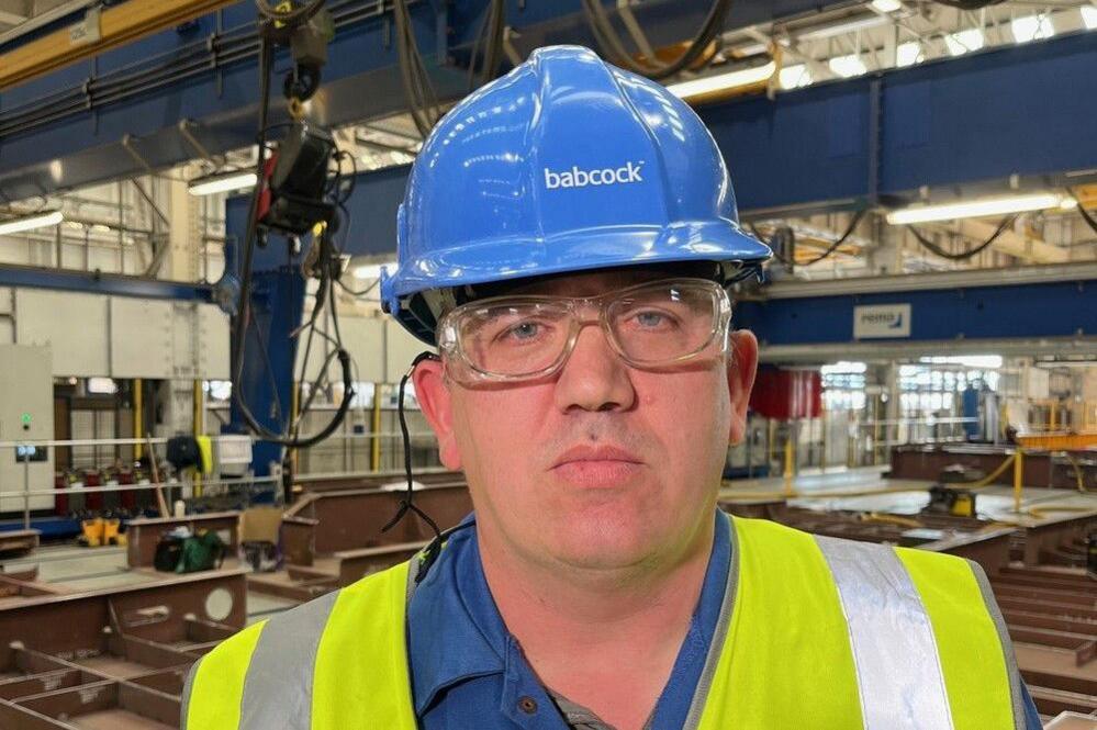 Paul Mclelland wears a white hard hat with the word babcock on it, and protective glasses. He stands inside a shipyard environment. He is wearing a blue jacket with a neon yellow high vis jacket over it.