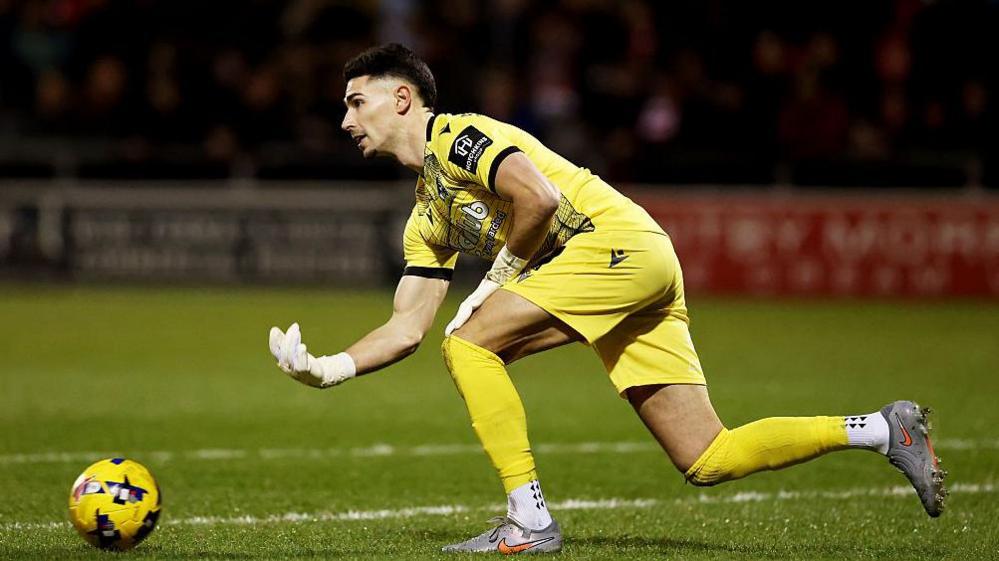 Luke Southwood, wearing an all-yellow kit, bowls the ball out along the ground for Bristol Rovers