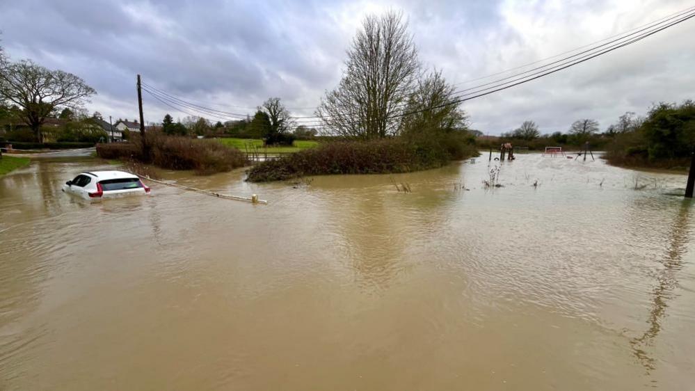 A white car is stuck in flood water. The water is above the tyres. A large area is covered by the water and houses can be seen in the background