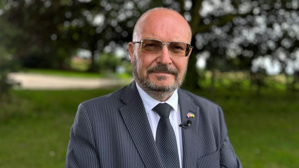 Graham Plant, dressed in a pinstripe suit, with black tie and white shirt. He is wearing tinted glasses, and has a full beard and moustache and close cropped hair. He is standing in parkland, with grass and mature trees in the background.