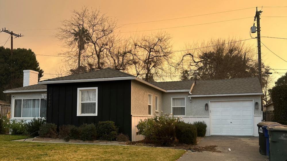 A bungalow on a street in America. The property has a short concrete driveway and a white garage. There is a small patch of grass in front of the house. The sky is a pale yellow and orange. The outline of the trees can be seen behind the house.