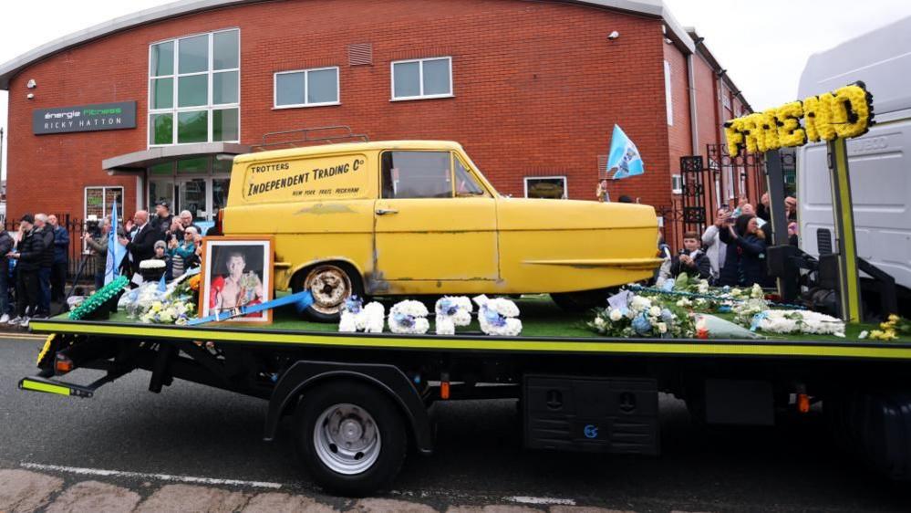 A little yellow three-wheeled van with a sticker on the back that reads 'Trotters Independent Trading Co' on a flatbed truck with is surrounded by flowers and a framed photo of Ricky Hatton.