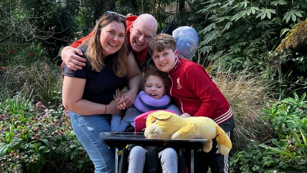 Addy poses for a family photo with her mother, father and brother in front of plants and flowers. She is in a specialist wheelchair.