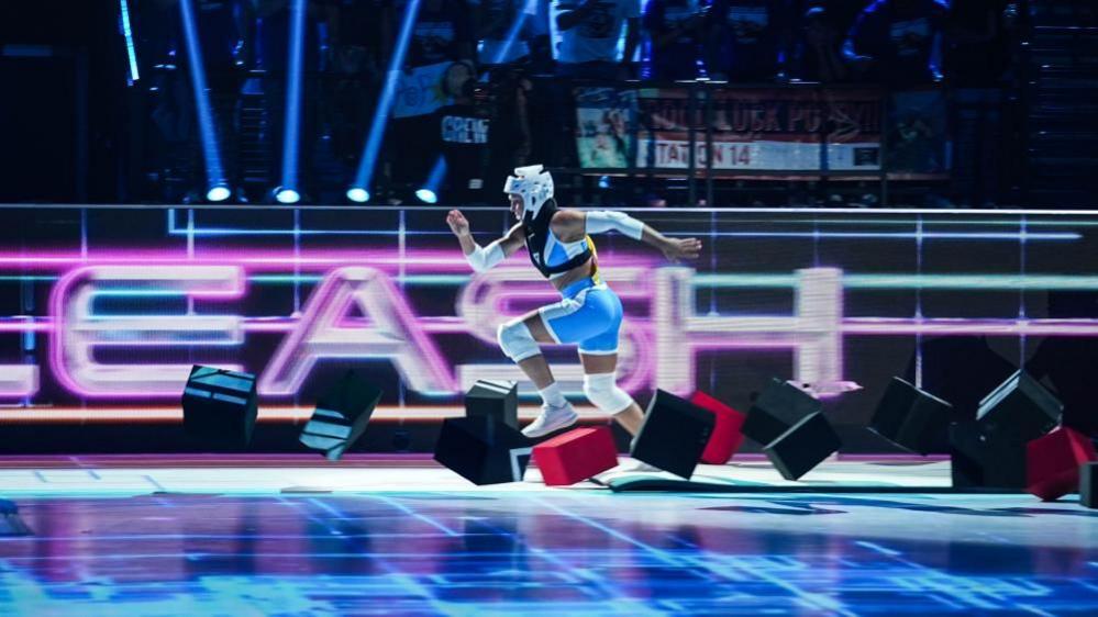 A female Gladiators contestant sprints through a colourful obstacle course. She wears blue shorts and a crop top with white helmet and knee pads. Neon lights in the background spell out the name of the challenge, 'Unleashed.'