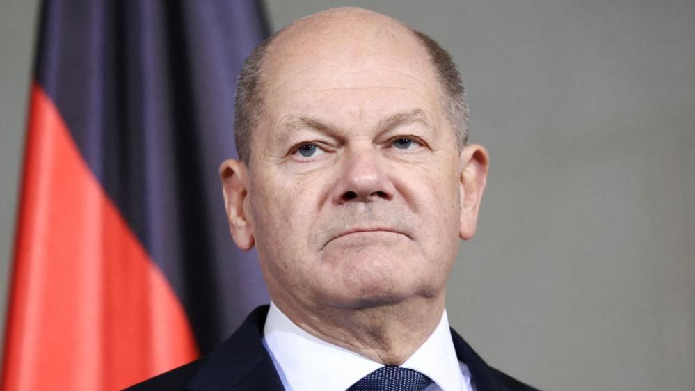 A headshot of Germany's Chancellor Olaf Scholz as he appears expressionless photographed in front of a red and black flag