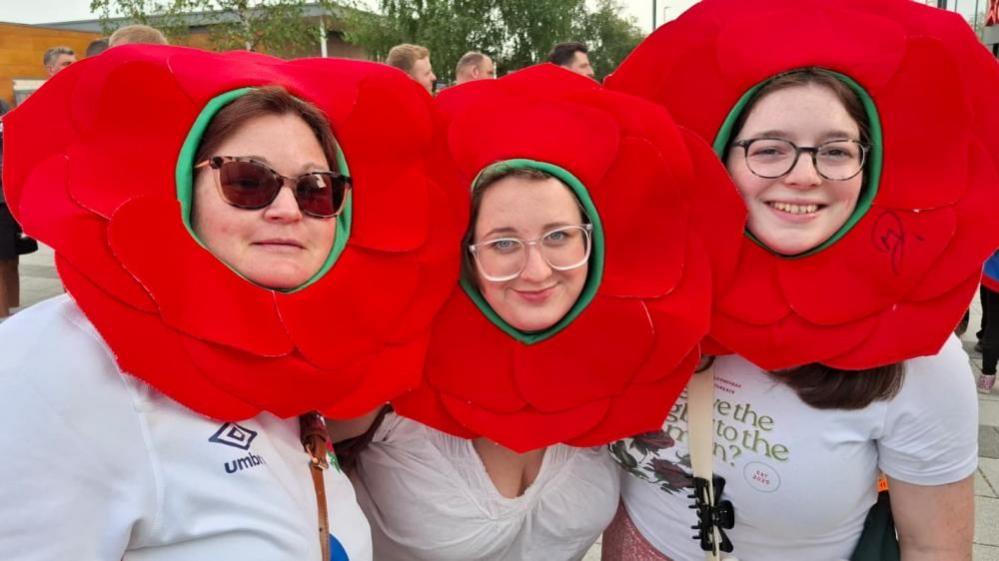 Three women look at the camera, smiling. They are wearing rose-shaped hats. Other people are behind them in a queue and all three have white tops on and glasses.