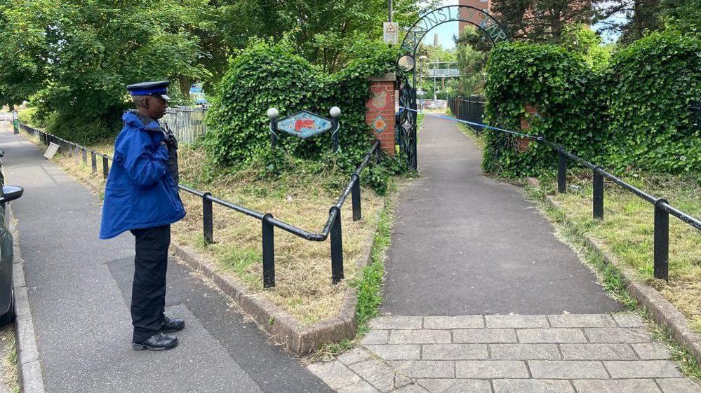 Entrance to a park - with wrought iron archway - cordoned off, with officer keeping watch