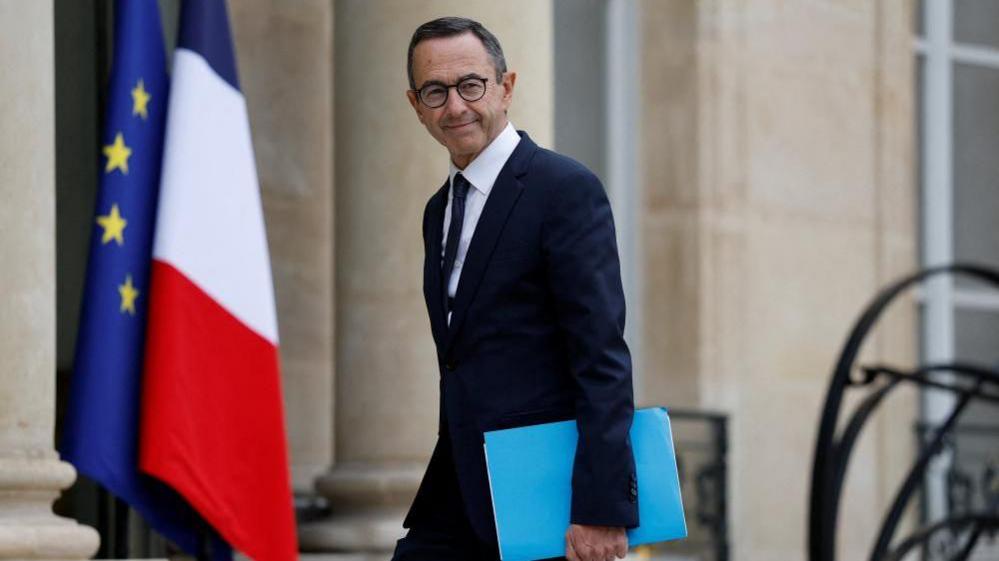 Bruno Retailleau, outgoing French Interior Minister, is pictured outside the Elysee Palace in front of French and EU flags. He is a man with dark hair in a dark suit wearing glasses and holding a blue folder.
