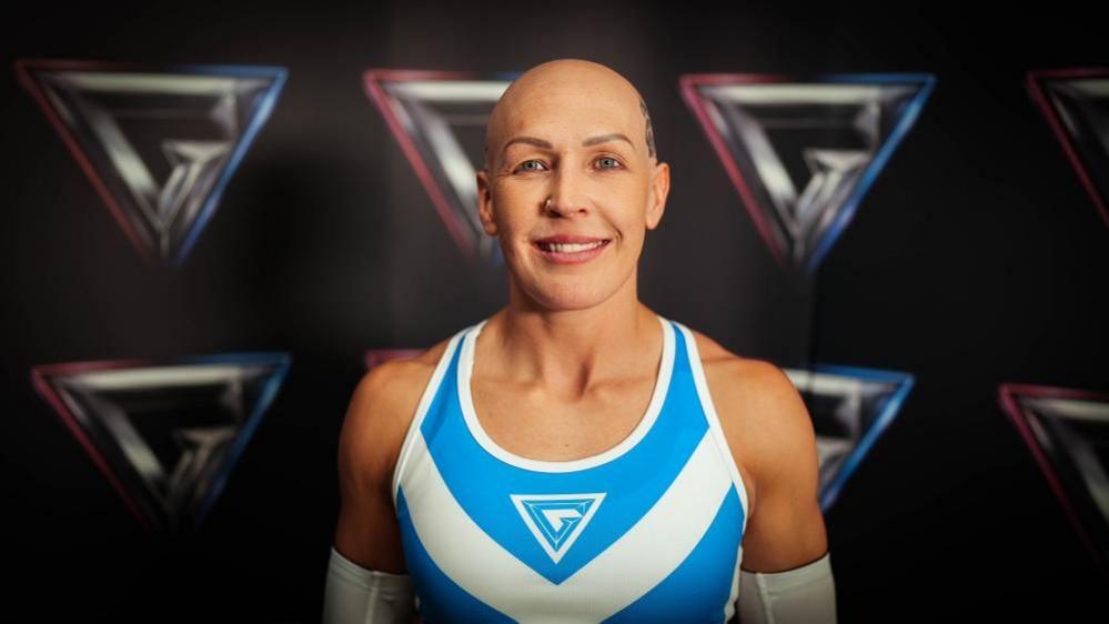 A white, female competitor wears the official 'blue team' kit and smiles happily in front of a Gladiator-branded backdrop.  She has a bald head, wears a little makeup and looks fit and muscular.