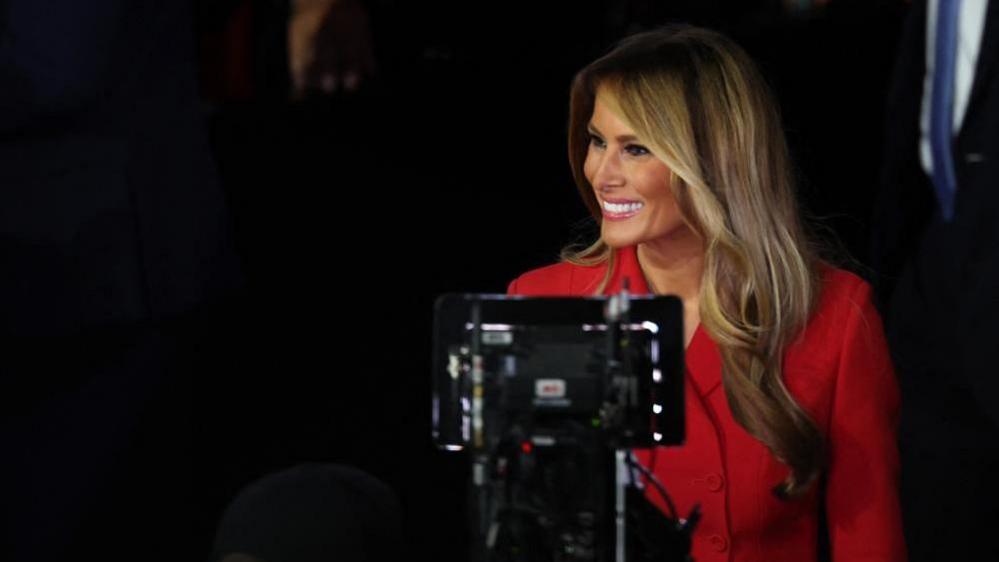 Melania Trump looks away from the camera, smiling. Video recording equipment is positioned in front of her. She is wearing a red jacket.