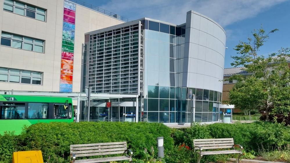 A large glass-fronted building with two park benches at its front. There is a green bus driving pass the entrance of the hospital. 