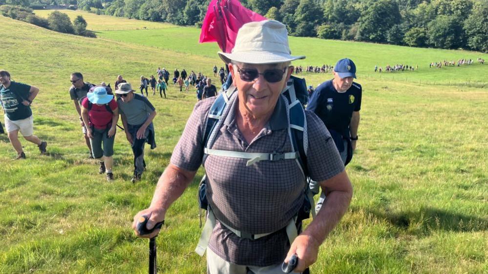 A man in his 70s smiles at the camera, with a long line of people walking behind him. He is wearing a sunhat and sunglasses, with walking poles in his hands.