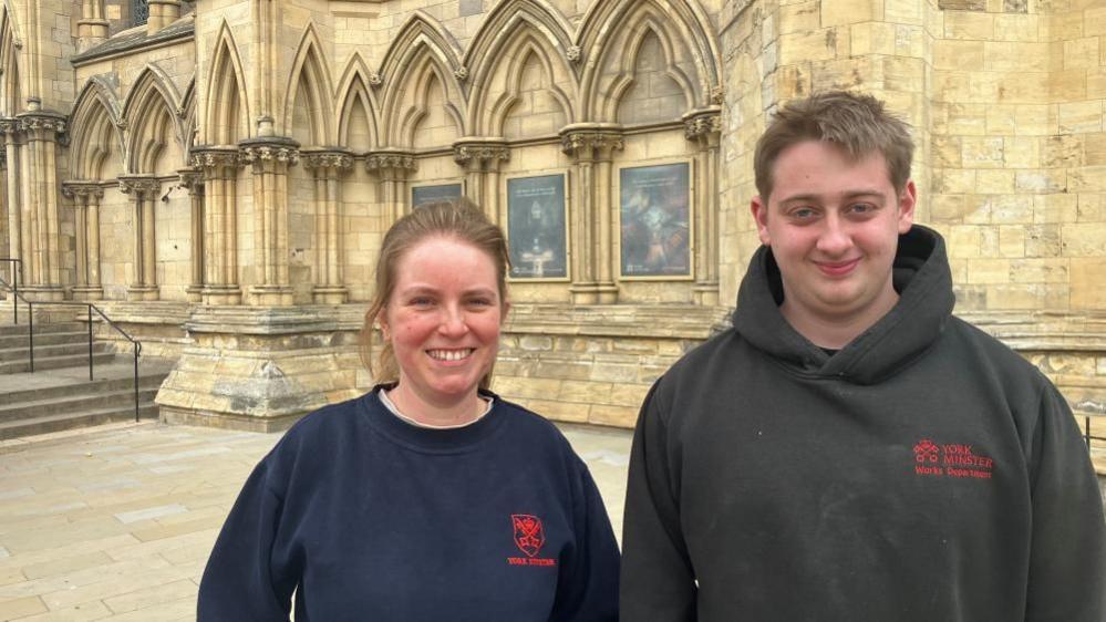 A young man and woman stand outside a historical building, wearing sweaters with the York Minster logo on the chest.