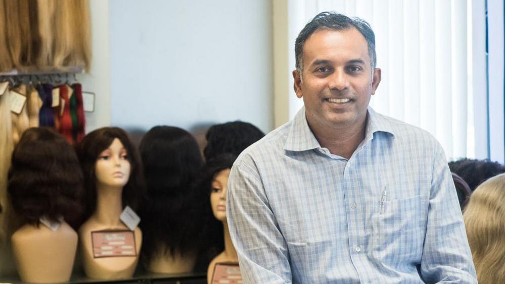 George Cherion smiling sits in front of mannequins wearing wigs. 