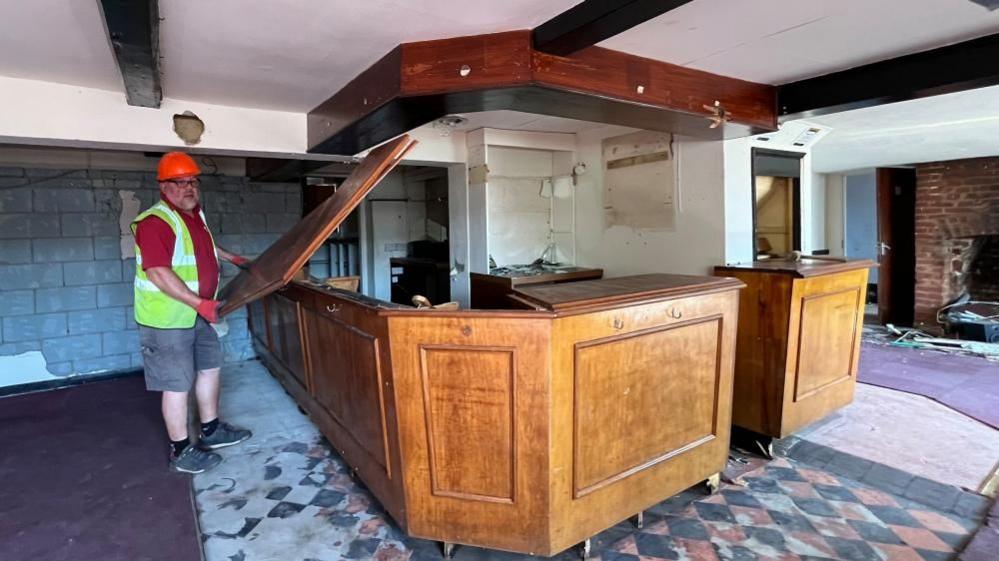 A salvage contractor removes the counter top of the bar of the Decoy Tavern. The plywood facade, with square trim panels, remains in situ, with an overbar on the ceiling and the floor tiles visible where the carpet has been removed.