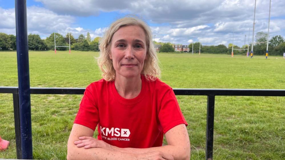 Katrina Collins sitting down at a table in the rugby club in a red T-shirt, with her arms crossed in front her.