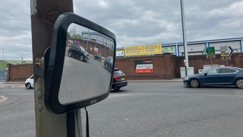 A convex mirror strapped to a lamppost on a traffic island at the junction of Lady Bay Bridge and Meadow Lane. Cars can be seen in the reflection of the mirror.