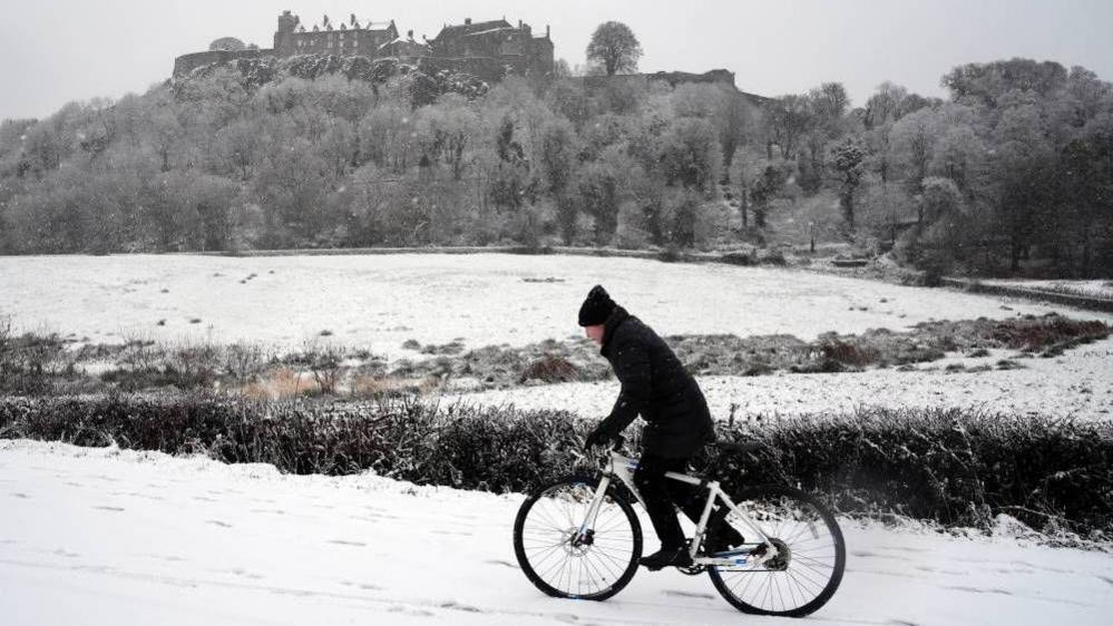 A man wearing a black coat and hat riding a bike through the snow. In the distance you can see a snowy field and Stirling Castle.