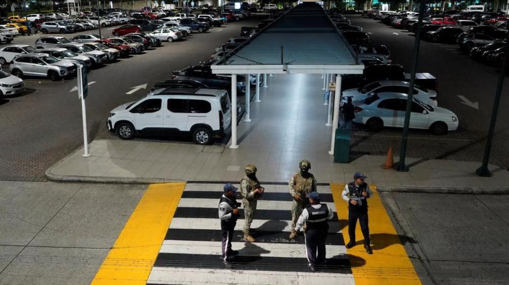 Members of Ecuador's armed forces stand stand guard after an armed attack at the airport in Guayaquil. There are two soldiers with guns and three other uniformed men standing on a pedestrian crossing leading to the parking lot.
