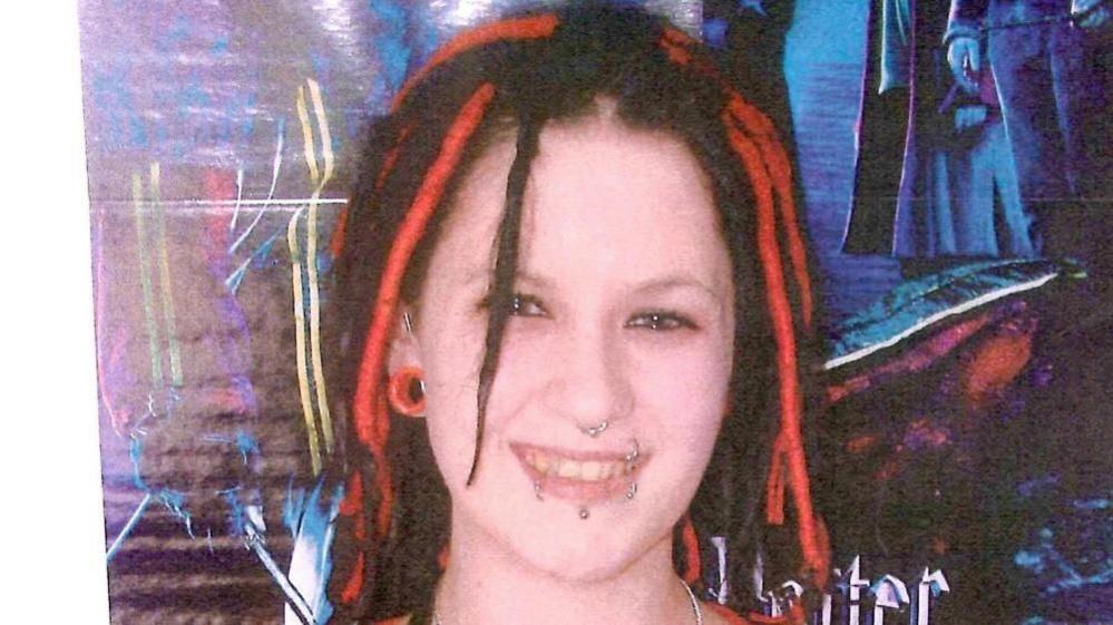 A photograph of a young woman smiling, stood against a graffiti-style backdrop. She has red and black dreadlocks, multiple lip piercings, a nose ring and a large stretcher-style earring.