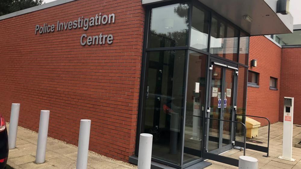 This is a picture of the Martlesham police investigation centre. On one side of the building, in grey letters against red bricks, it spells out  police investigation centre . There are automatic sliding doors at the front of the building  and a free standing intercom visible. There are also white bollards running alongside the building. 